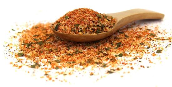 Ein Holzlöffel, gefüllt mit einer Gewürzmischung aus getrockneten Kräutern und roten Gewürzen, die auf einer weißen Oberfläche verstreut sind.