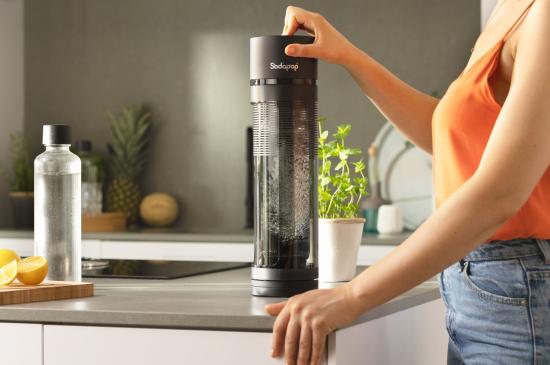 Person bedient einen Sodastream auf einer Küchenarbeitsplatte, umgeben von Obst und Kräutern.