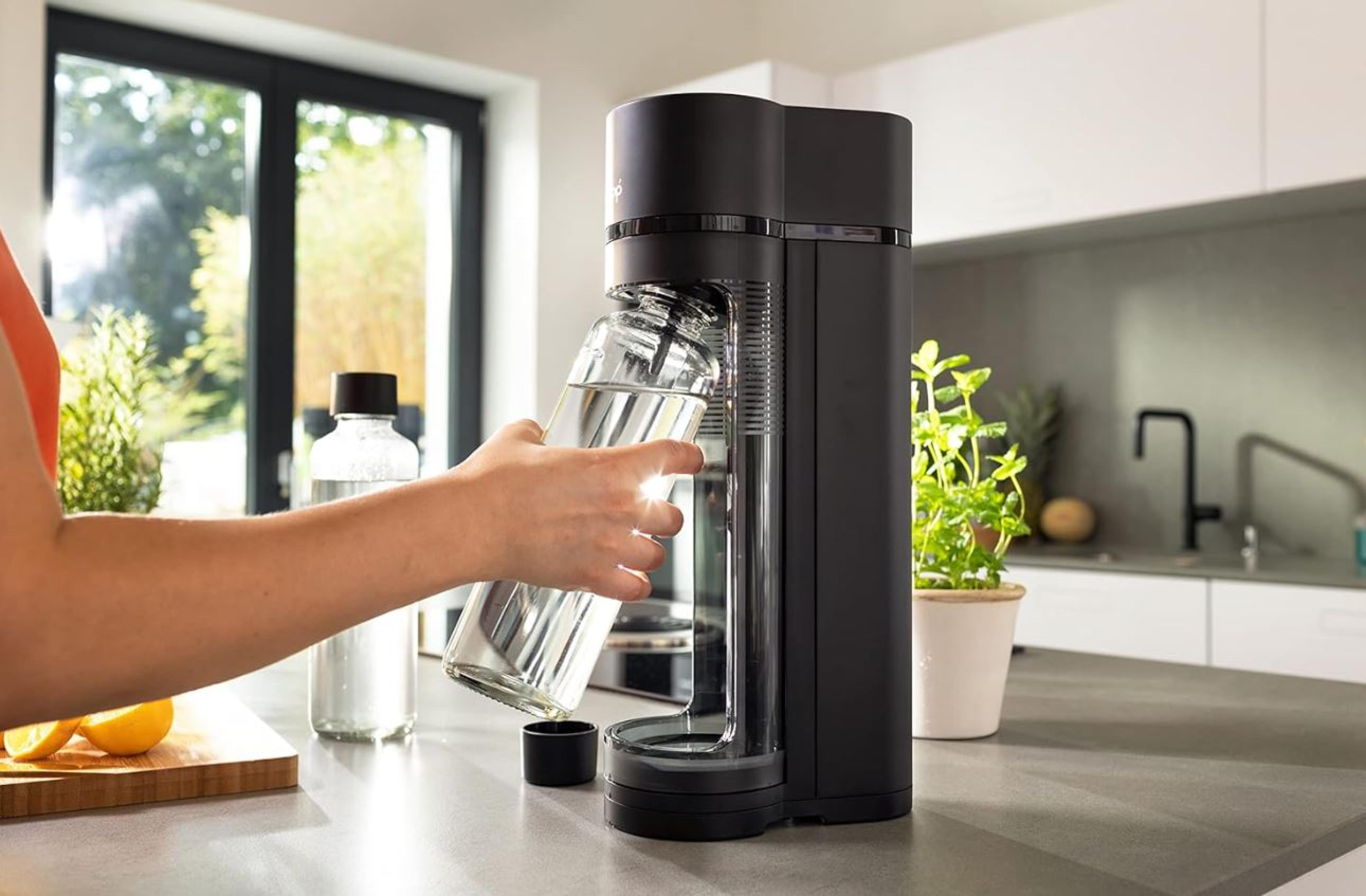 Eine Person füllt eine Glasflasche mit Sprudelwasser aus einer schwarzen Soda-Maschine in einer modernen Küche