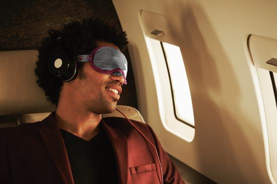 Mann im Flugzeug mit Schlafmaske und Kopfhörern, lächelnd auf seinem Sitz