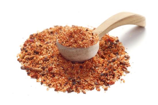 Ein Holzlöffel, gefüllt mit einer roten Gewürzmischung, liegt auf einem Haufen derselben Würzmischung auf einem weißen Hintergrund.