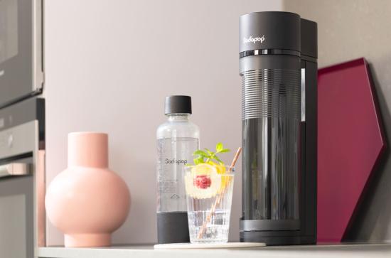 Ein schwarzer Wassersprudler steht neben einer Wasserflasche und einem Glas mit Getränk und Zitrusfrüchten auf einer Küchenarbeitsplatte.